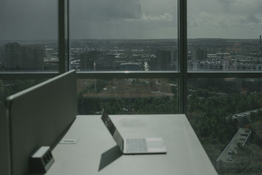 Laptop on office desk with a city skyline through a large window, offering a modern workspace view.